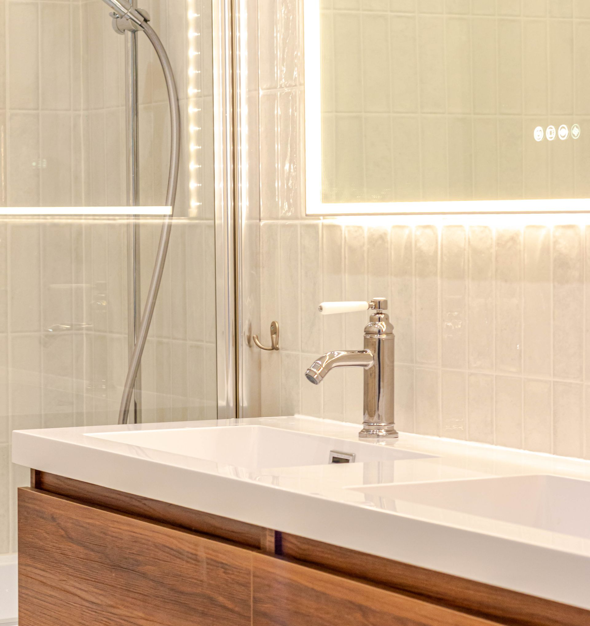 Lavabo de salle de bain avec robinet chromé, comptoir blanc, armoire en bois et douche derrière verre.
