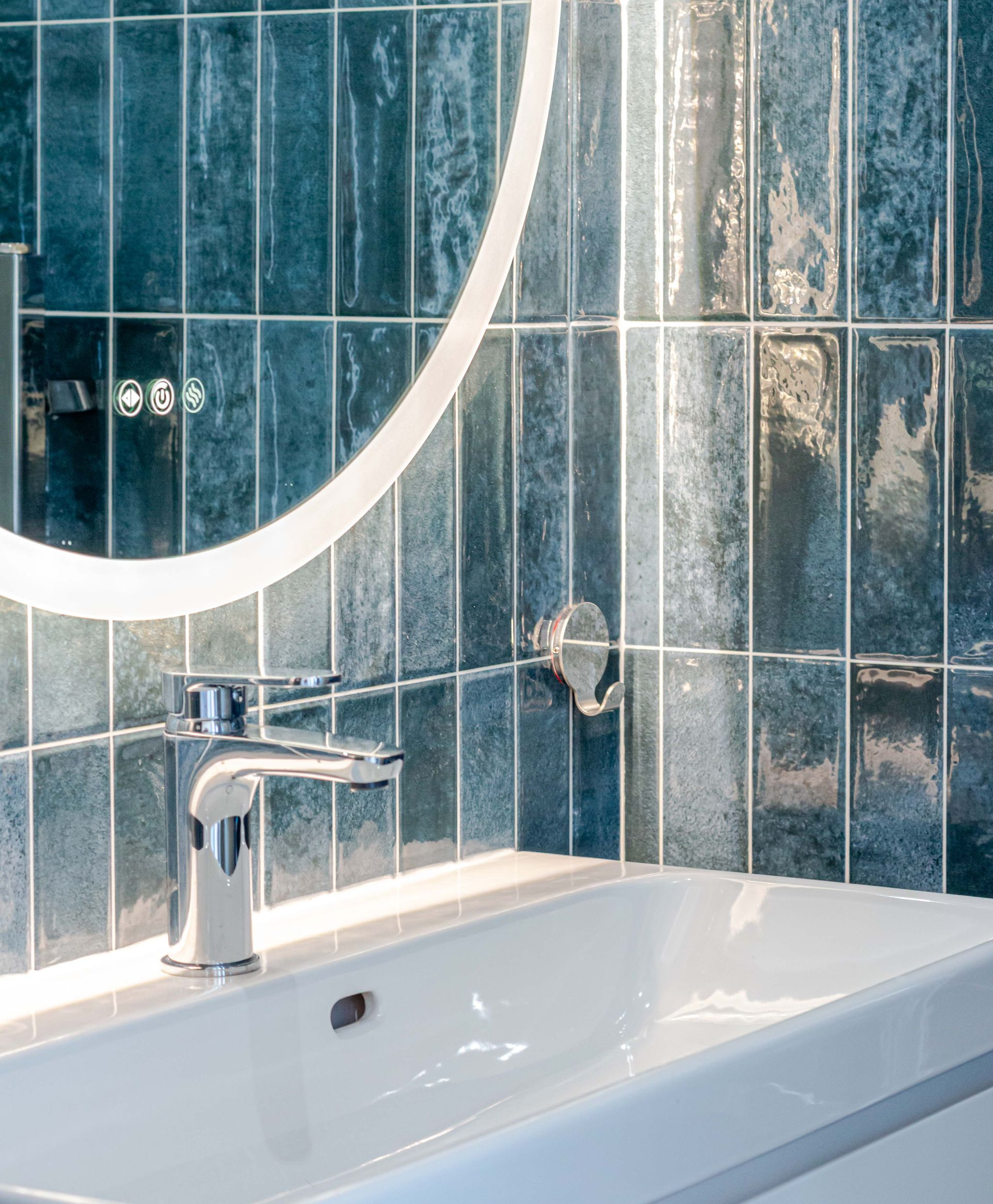 Lavabo blanc avec robinet chromé et carreaux verticaux bleus, miroir éclairé et crochet porte-serviettes.