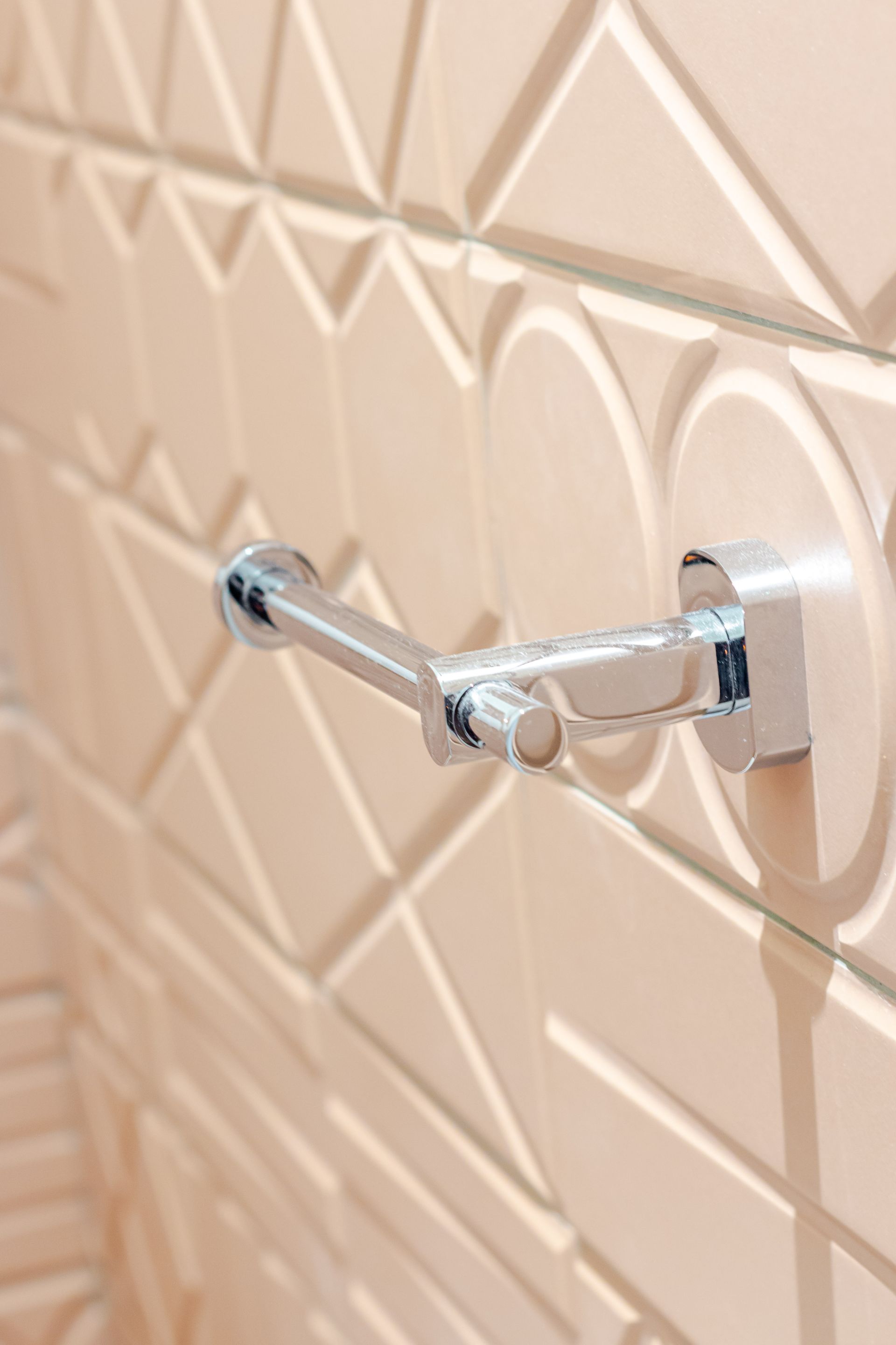 Porte-papier toilette chromé monté sur un mur en carrelage géométrique texturé de couleur pêche.