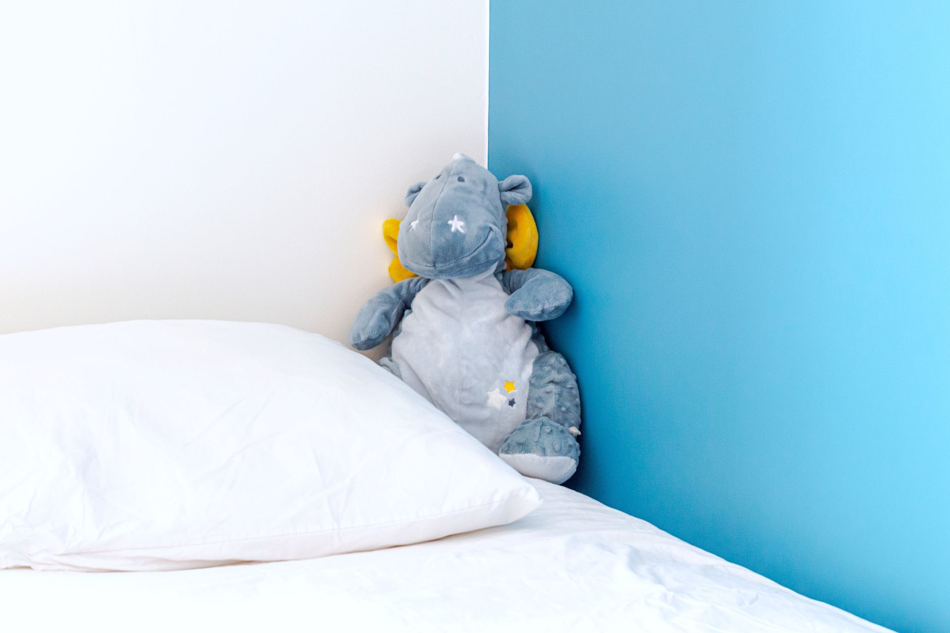 Un hippopotame en peluche est assis dans le coin d'un lit avec un oreiller blanc et des murs bleus et blancs.