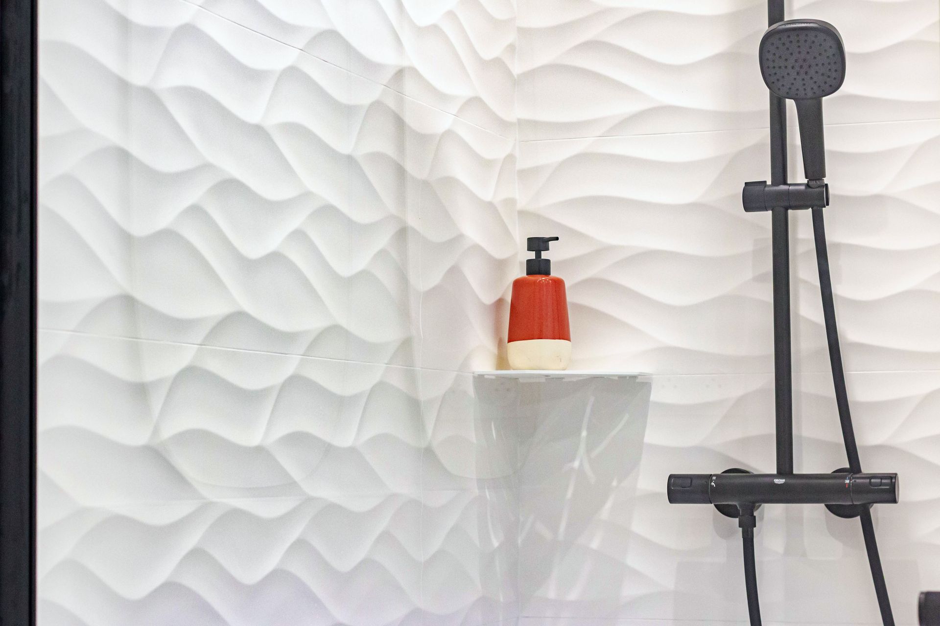Mur de douche en carrelage blanc à motifs ondulés avec une étagère sur laquelle est fixé un distributeur de savon.