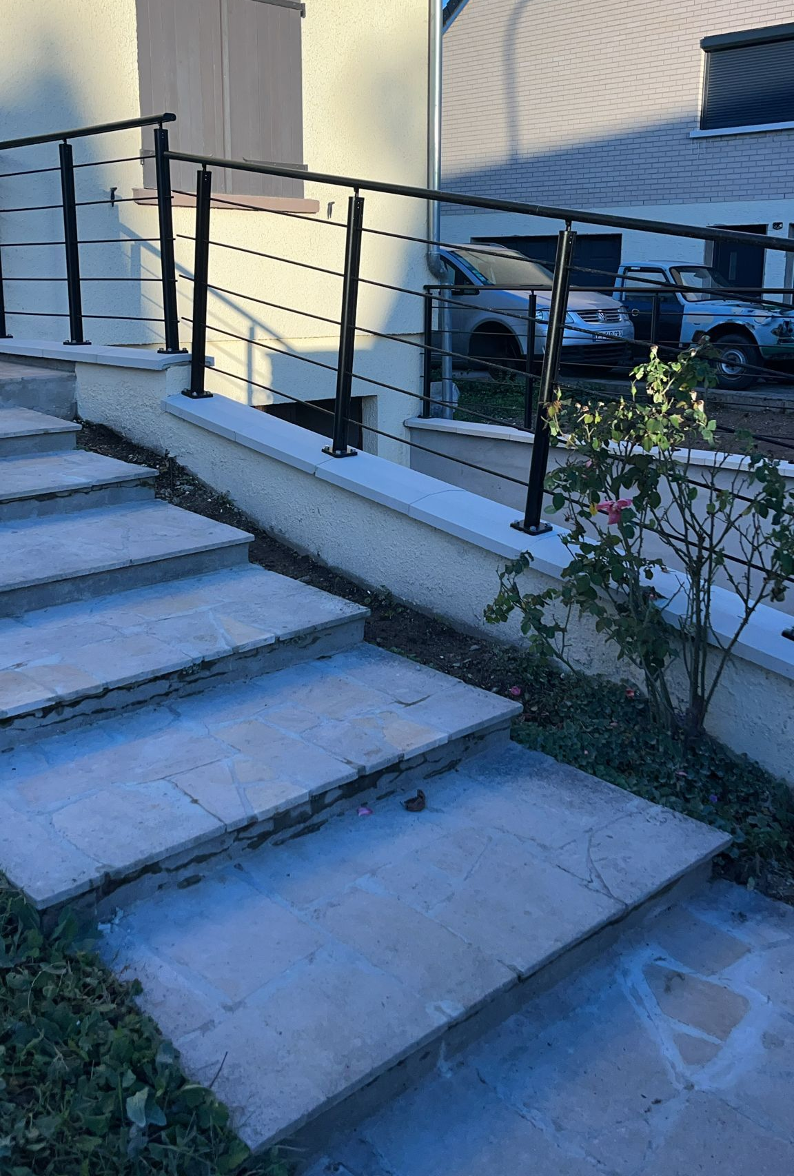 Un escalier en pierre mène à un bâtiment avec une rampe en métal noir. Une voiture est visible en arrière-plan.