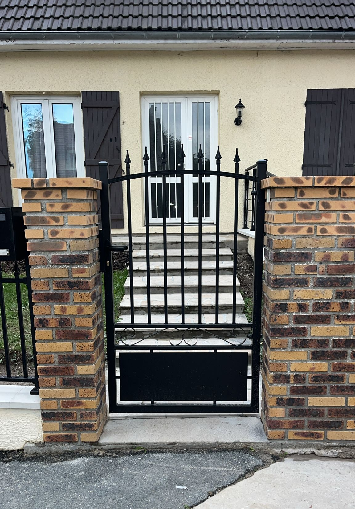 Portail en fer forgé noir entre des piliers en briques menant à une porte blanche.