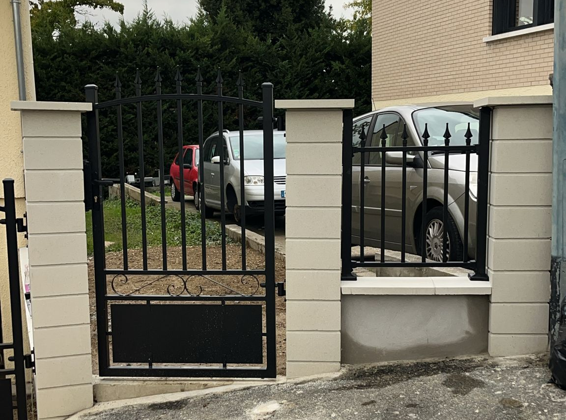 Portail et clôture en fer forgé noir sur piliers en briques beige clair ; voitures garées derrière le portail.
