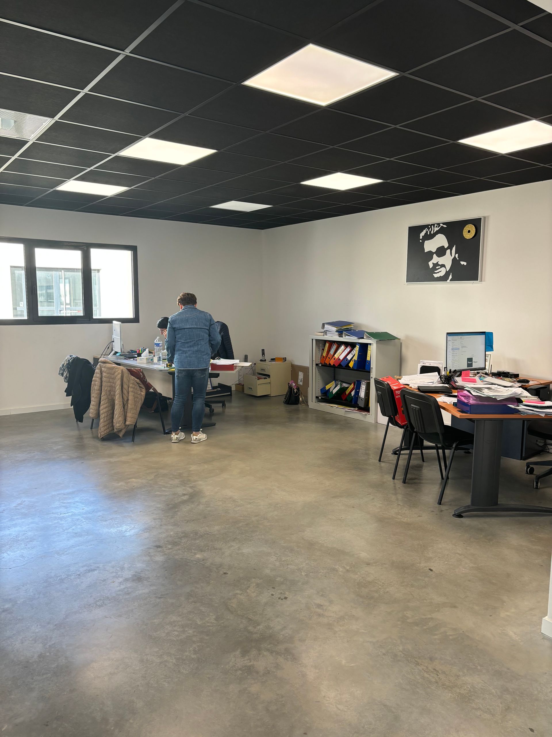 Une personne se tient debout à un bureau dans un bureau au plafond à grille noire, au sol en béton et avec une affiche au mur.