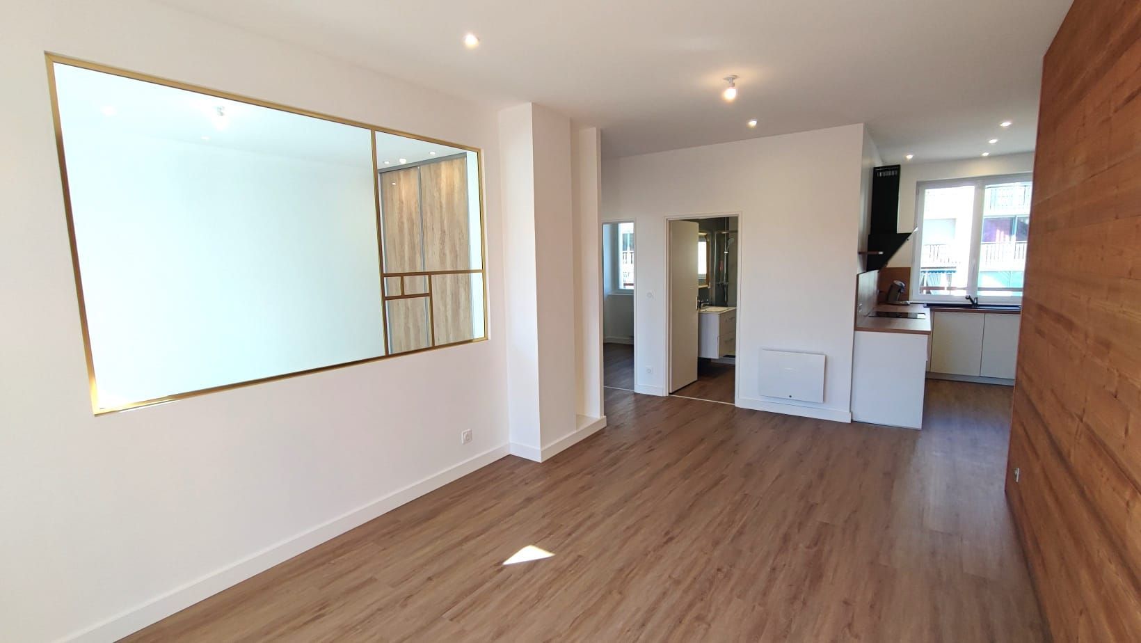 Intérieur d'appartement vide avec parquet, grand miroir et cuisine partiellement visible.