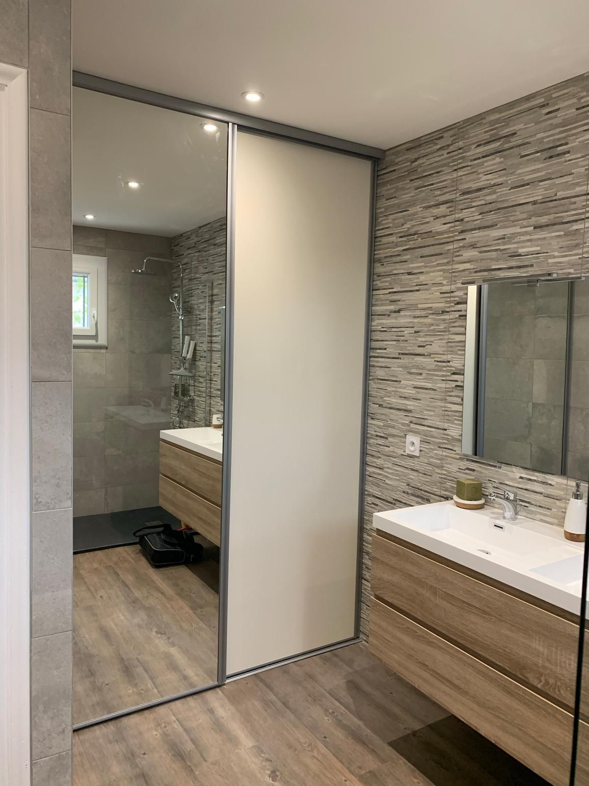 Salle de bains moderne avec meuble-lavabo en bois, mur imitation pierre et grande porte coulissante avec panneau miroir reflétant la douche.