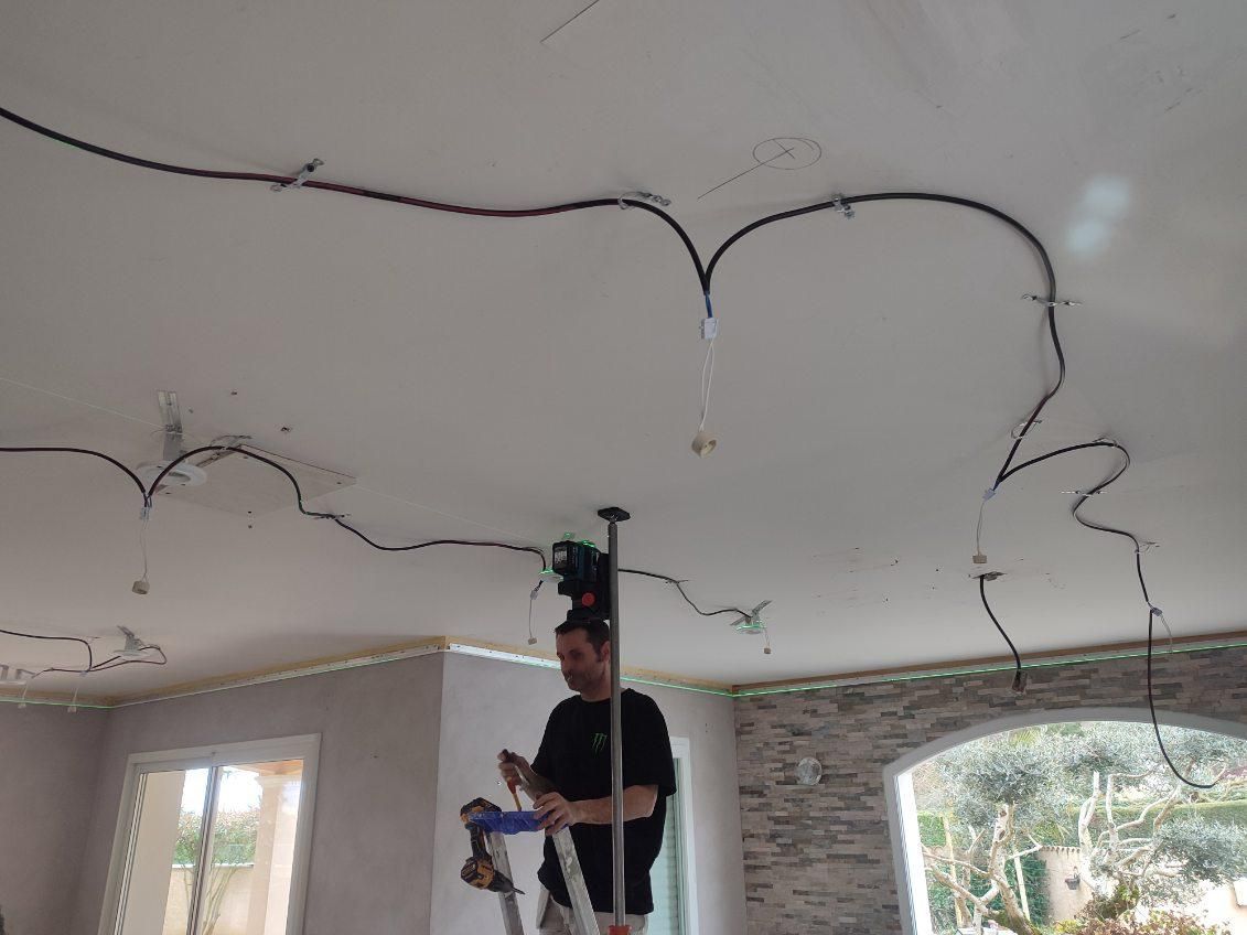 Un homme, sur une échelle, installe des câbles électriques sur un plafond blanc. Les fils sont fixés par des clips.