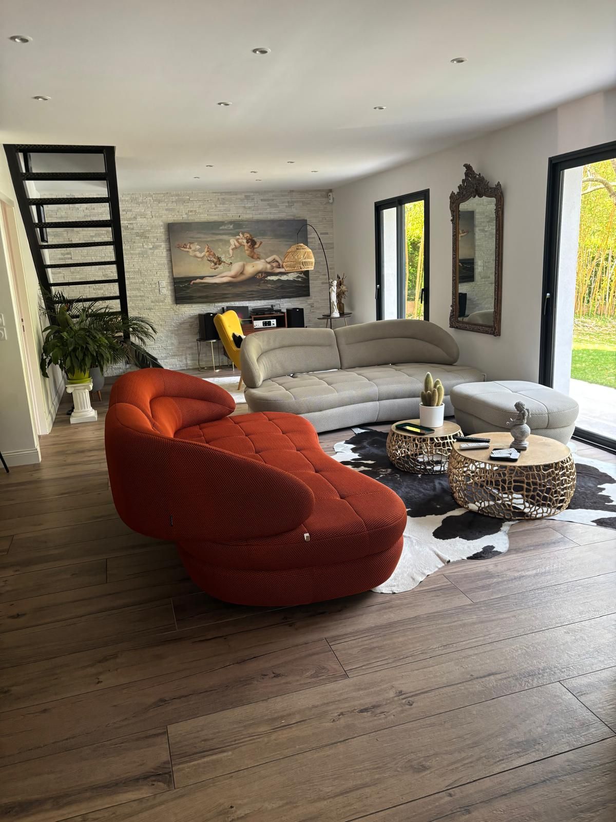Un salon moderne avec un canapé orange, un canapé gris incurvé, un tapis en peau de vache, des tables basses rondes et un escalier en métal.