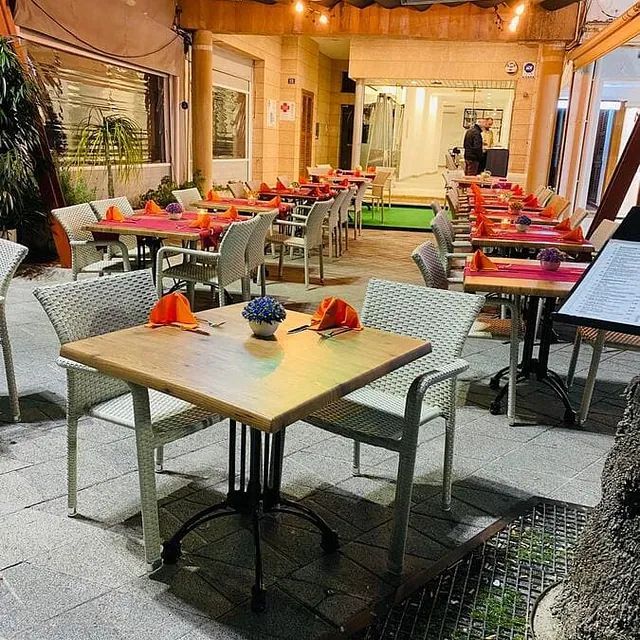 Patio de restaurante al aire libre con mesas vacías, sillas de mimbre y servilletas rojas bajo luces cálidas.