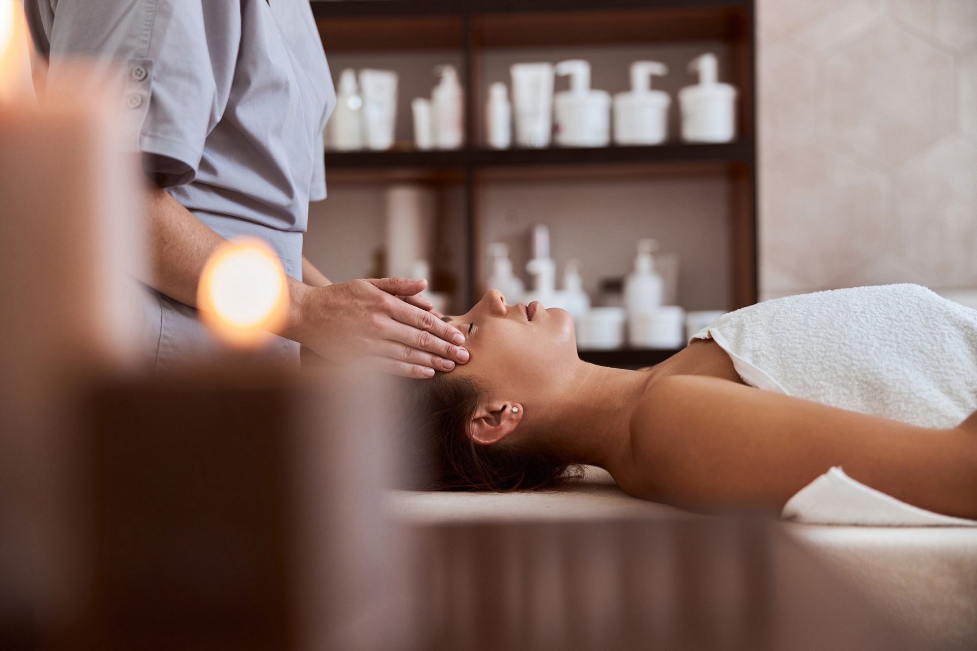 Mujer recibiendo un tratamiento facial en un spa. Las manos de la terapeuta sobre su rostro; velas encendidas en primer plano, estantes con productos.