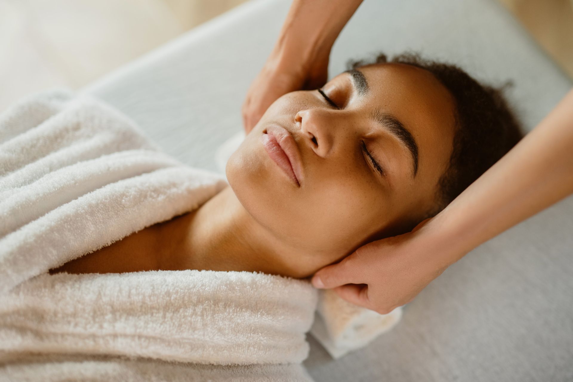 Persona recibiendo un masaje de cuello, con los ojos cerrados, tumbada en una mesa de masajes, vestida con una bata blanca.