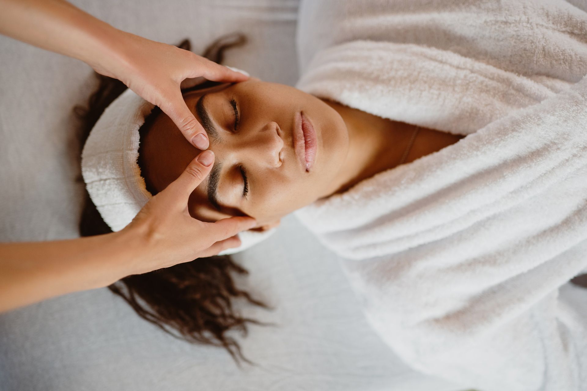 Mujer recibiendo un masaje facial en un spa. Con las manos en la frente, en bata y con los ojos cerrados.