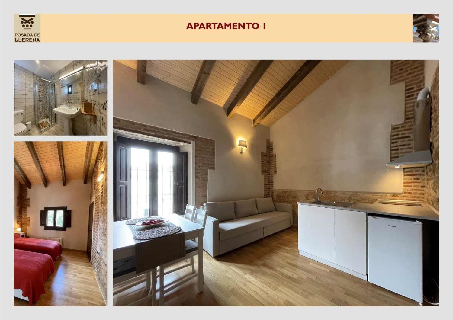 Acogedor estudio con vigas a la vista, sofá gris, cocina pequeña y alcoba roja para dormir; distribución en dos ambientes
