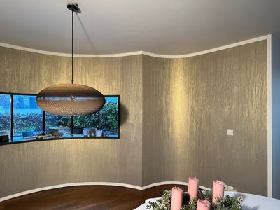 Eine mit Staub bedeckte Hand hält einen Pinsel und trägt einen weissen Anstrich auf eine raue, strukturierte Wand auf.