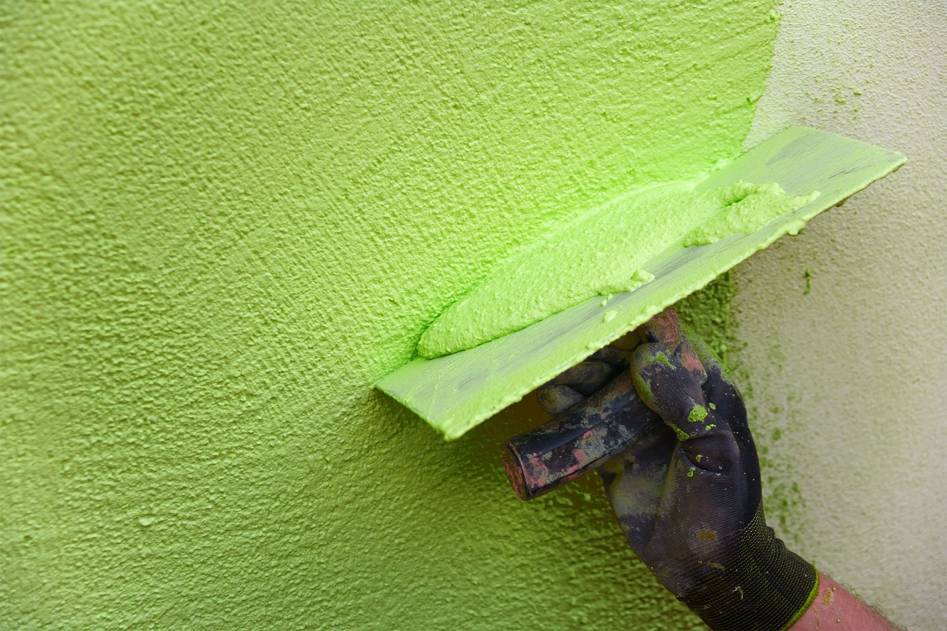 Eine behandschuhte Hand trägt mit einer Kelle hellgrünen Strukturputz auf eine Wand auf.
