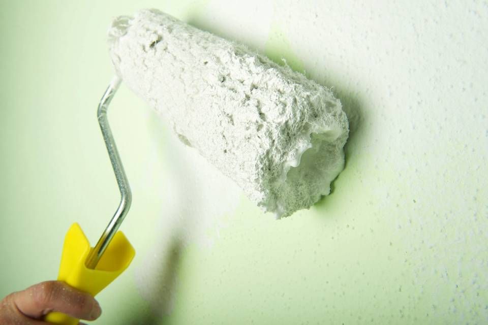 Eine Hand, die eine Farbrolle mit gelbem Griff hält, trägt dicke weisse Farbe auf eine hellgrüne Wand auf.