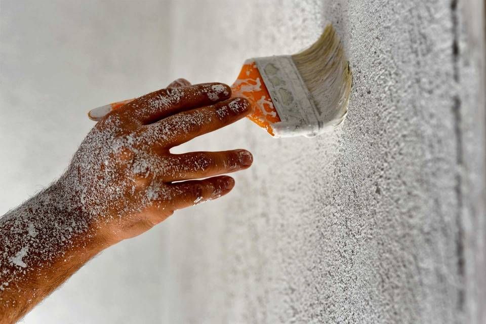Eine mit weissem Staub bedeckte Hand hält einen Pinsel und trägt einen weissen Anstrich auf eine raue, strukturierte Wand auf.