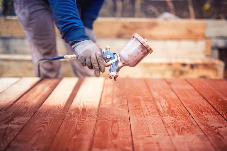 Eine Person in Arbeitshandschuhen trägt mit einer Spritzpistole eine rötlich-braune Holzbeize auf mehrere Holzplanken auf.