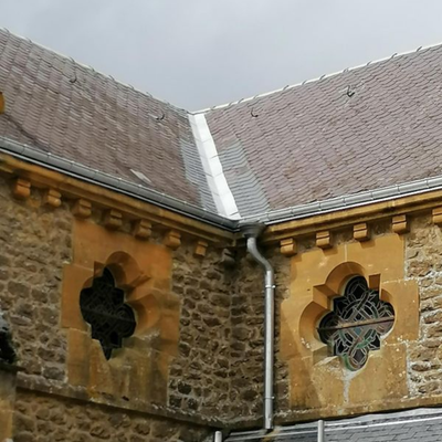 Angle d'un bâtiment en pierre avec deux fenêtres cintrées décoratives et un toit de tuiles pentu sous un ciel nuageux