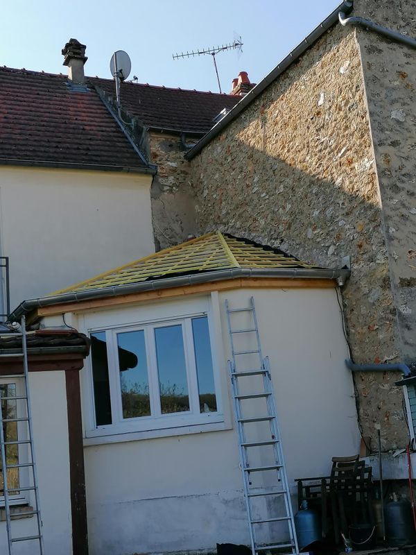 Extérieur de la maison avec une échelle appuyée contre un mur blanc et une petite extension au toit jaune.