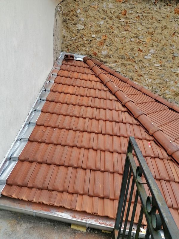 Toit étroit recouvert de tuiles en terre cuite rouge, jouxtant un mur blanc et une façade en pierre.