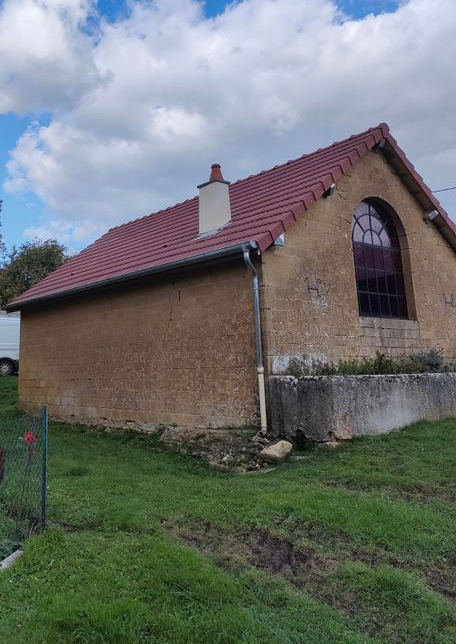 Petite maison en briques au toit rouge et à la fenêtre cintrée, vue de côté sous un ciel nuageux.