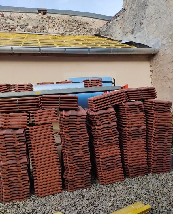 Des piles de tuiles en terre cuite sont entreposées dans une cour de gravier, à côté d'un mur de bâtiment.