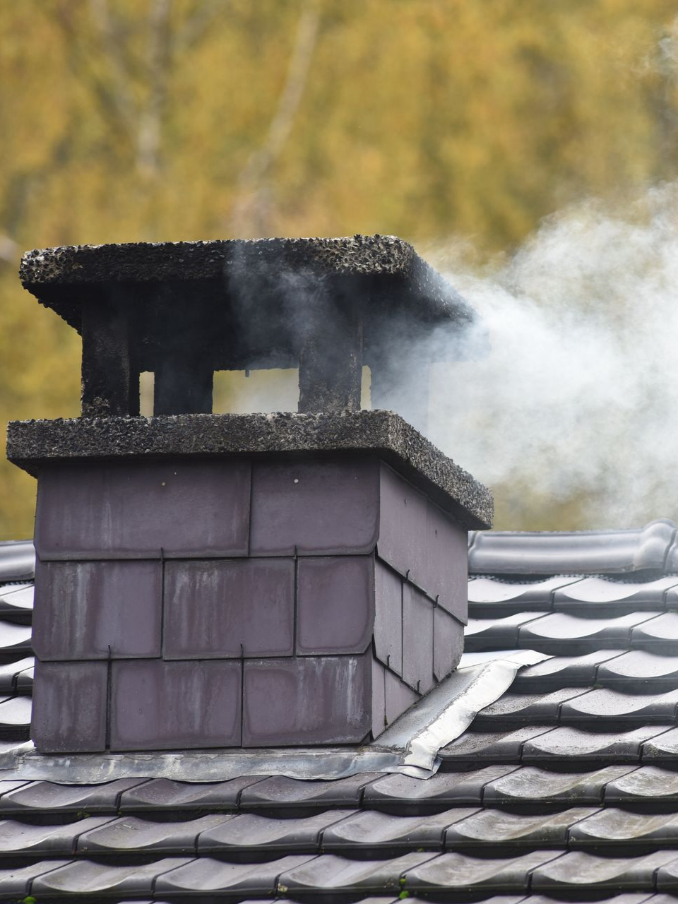 De la fumée s'échappe d'une cheminée sur le toit d'une maison aux bardeaux gris et au chapeau de pierre.