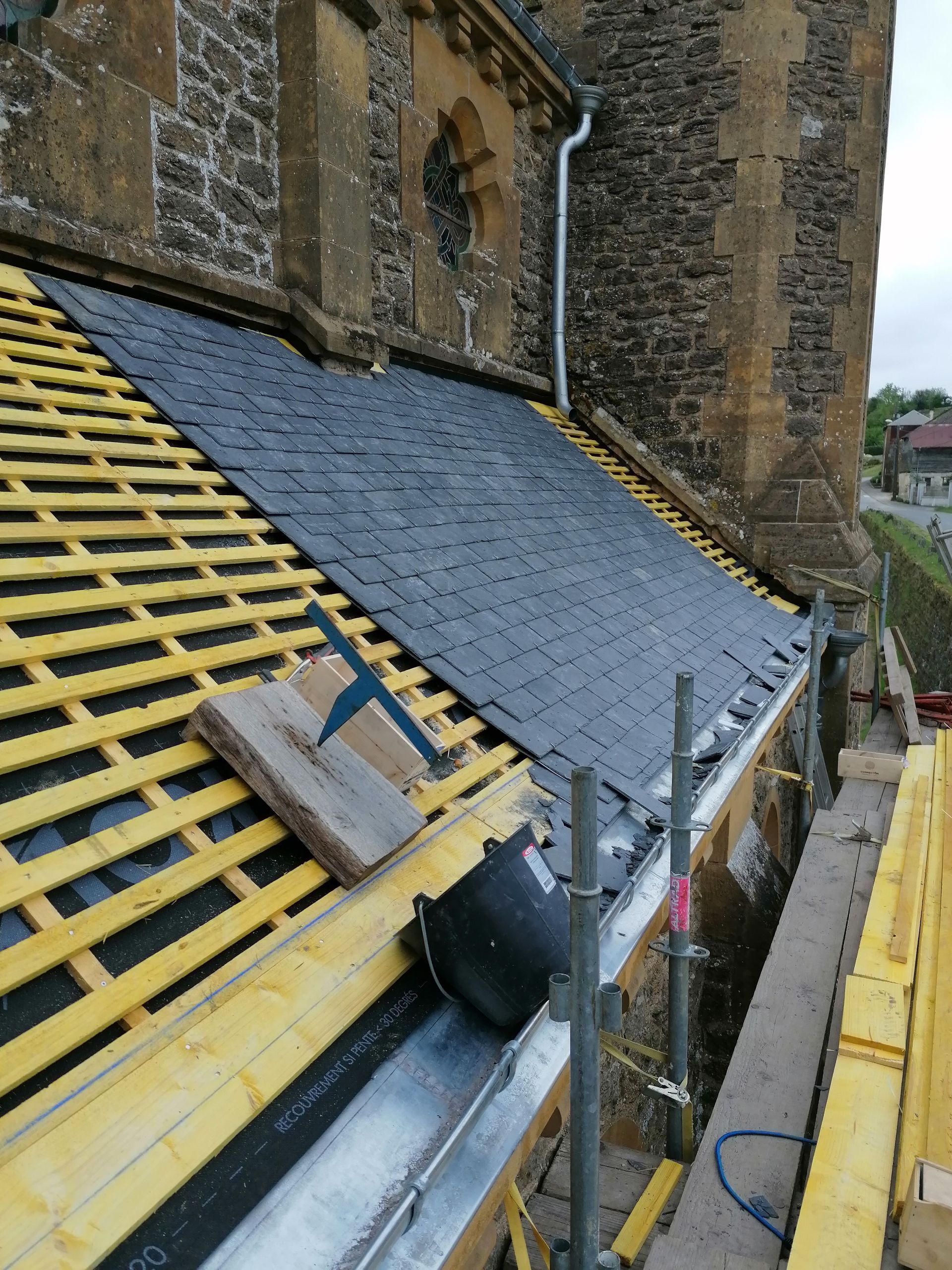 Travaux de réparation de toiture sur un toit d'ardoise à forte pente jouxtant un bâtiment en pierre, avec échafaudages et outils.
