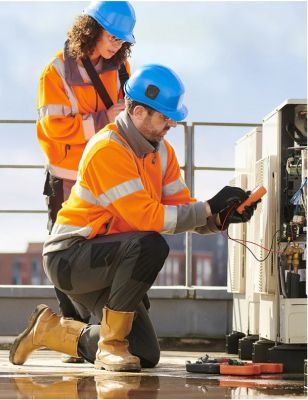Dos trabajadores con trajes de seguridad naranjas inspeccionan equipos eléctricos al aire libre.
