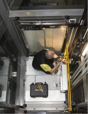 Técnico trabajando dentro de un hueco de ascensor junto a barandillas de seguridad amarillas, llevando una caja de herramientas y herramientas.