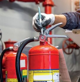 Mano enguantada utilizando una llave inglesa para dar mantenimiento a un extintor de incendios rojo en un entorno industrial.