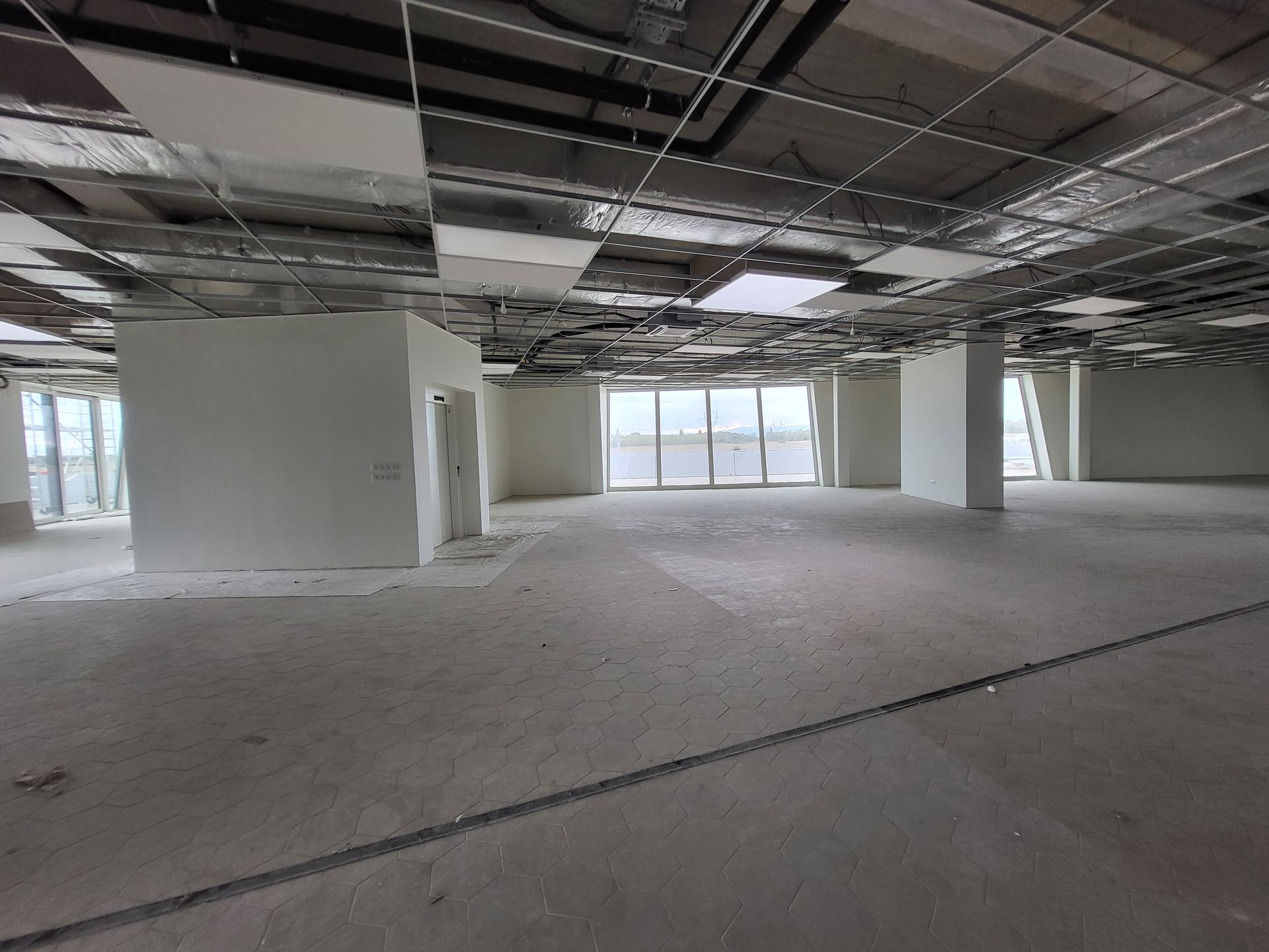 Espace de bureaux vide et inachevé, avec un plafond apparent, des murs blancs et de grandes fenêtres.