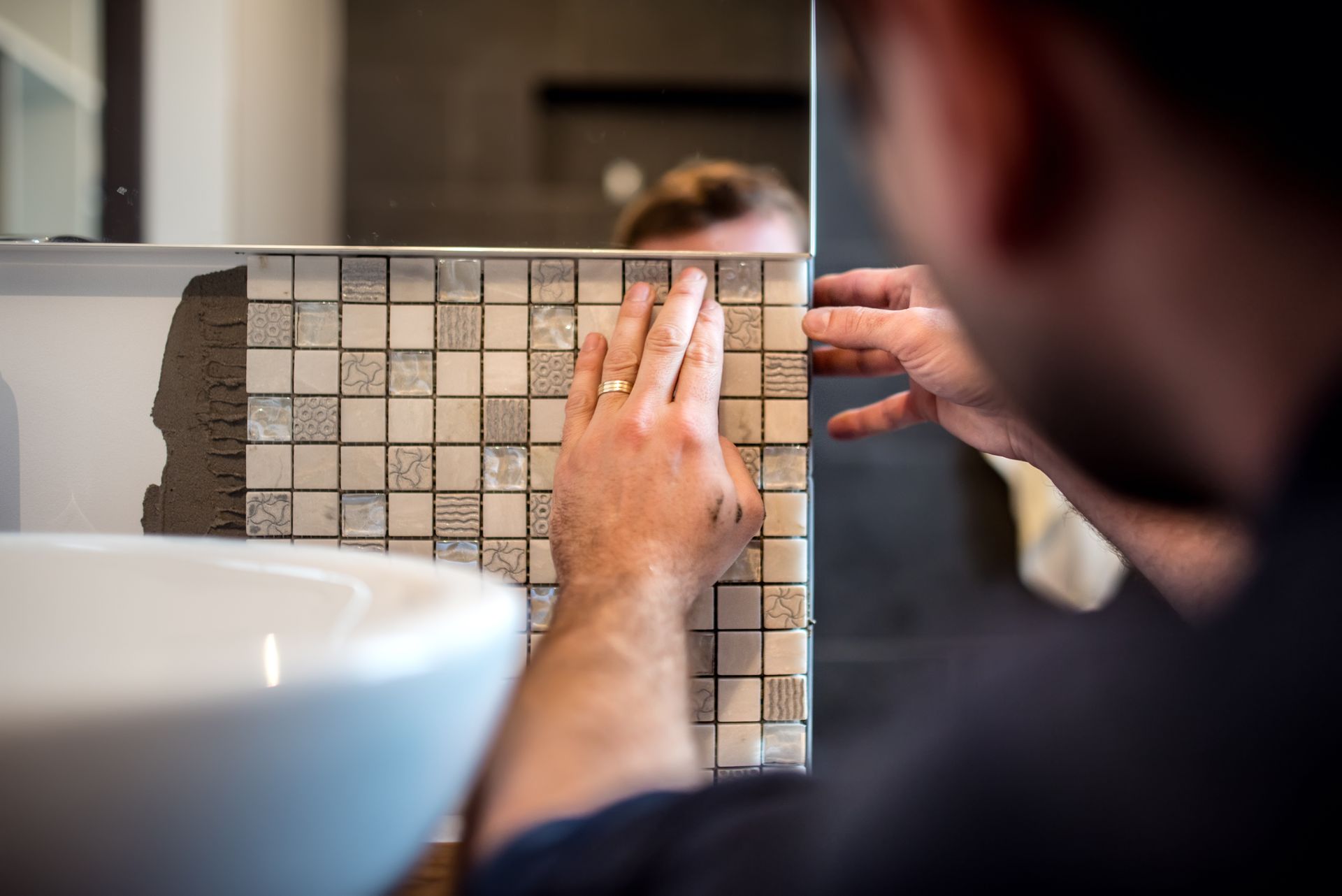 Une personne pose des mosaïques carrées beiges sur le mur d’une salle de bains, près d’un lavabo, en s’aidant d’un niveau.