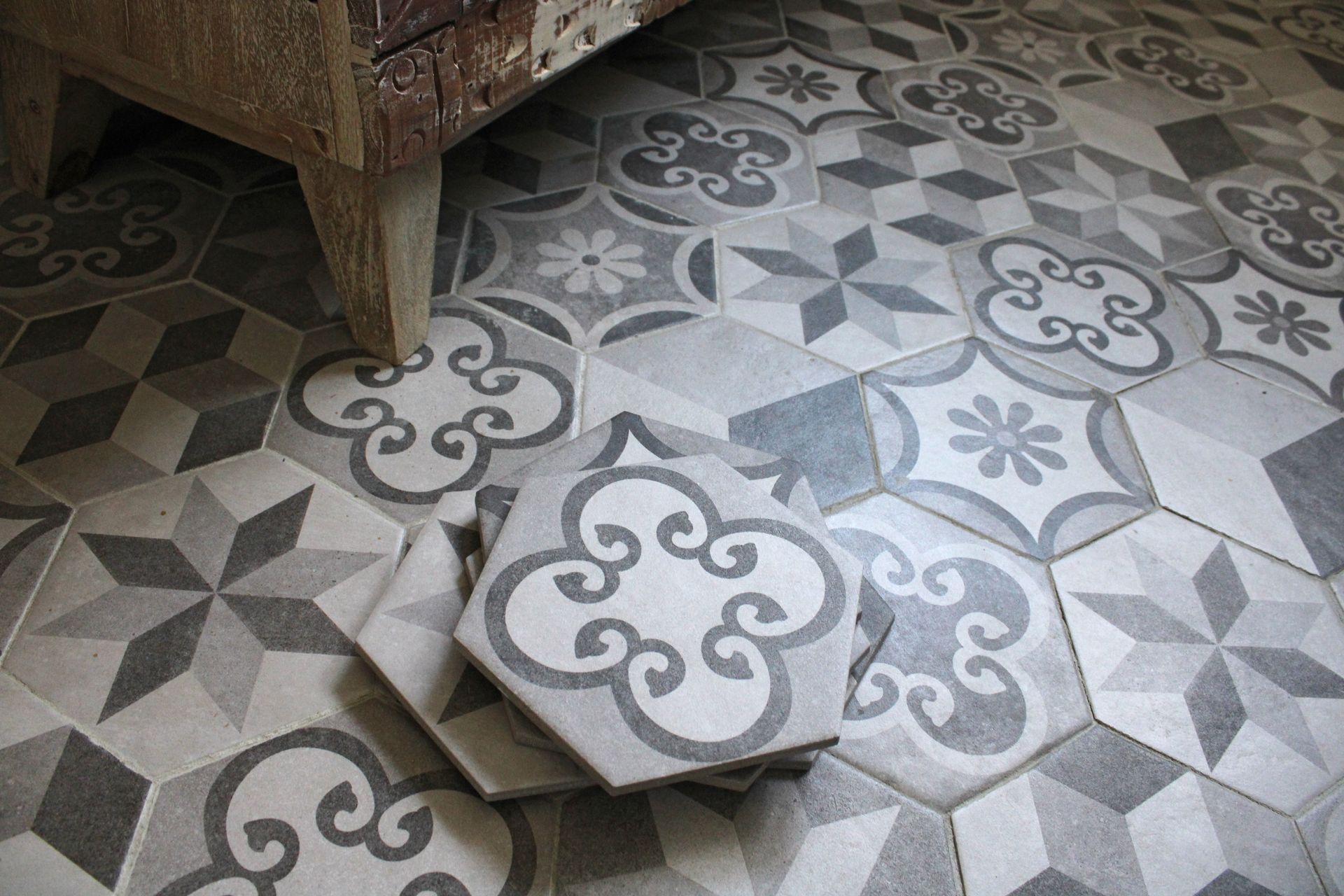 Carreaux de sol gris et blancs à motifs, avec un carreau décollé près d’un pied de chaise en bois.