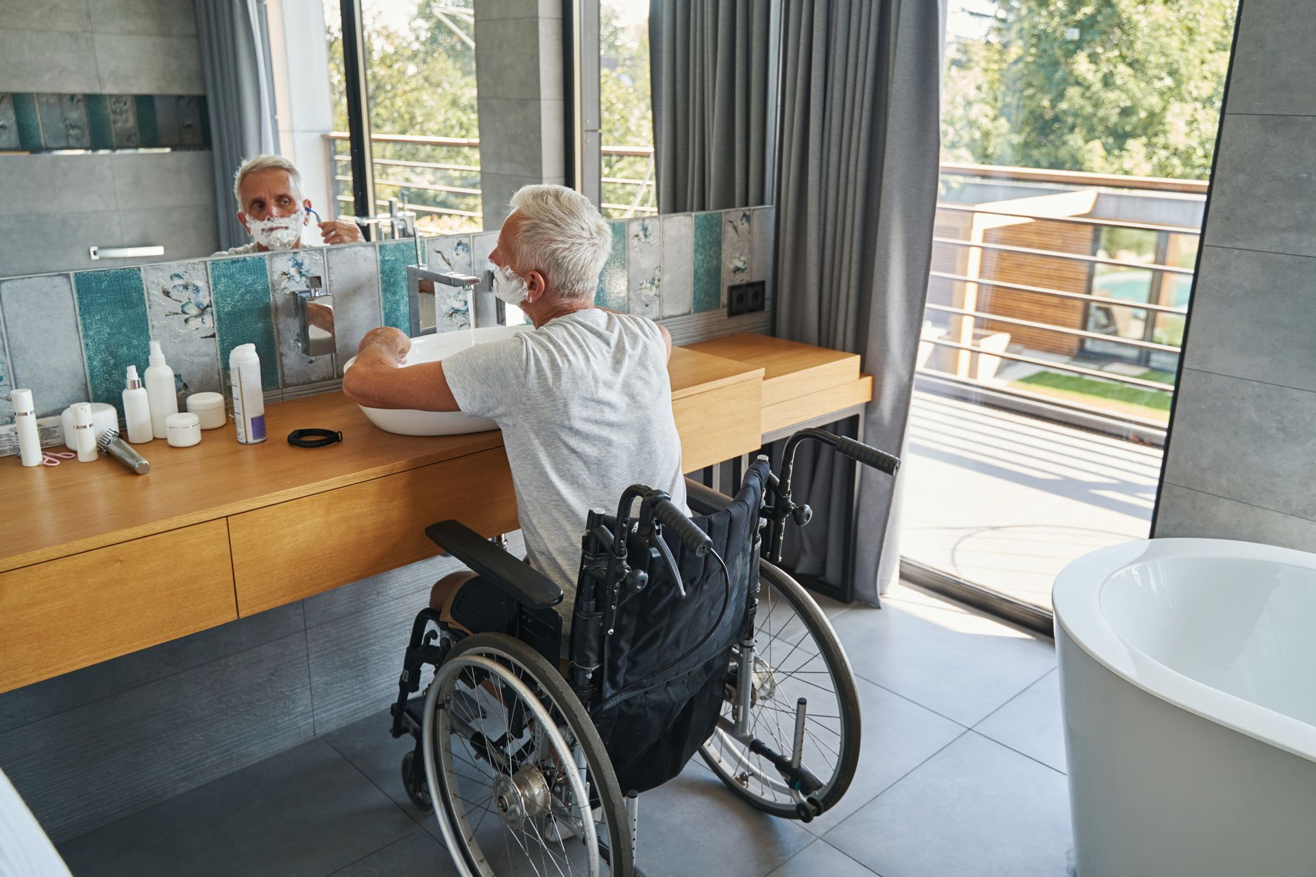 Une personne en fauteuil roulant se rase au-dessus du lavabo d’une salle de bains moderne et lumineuse.