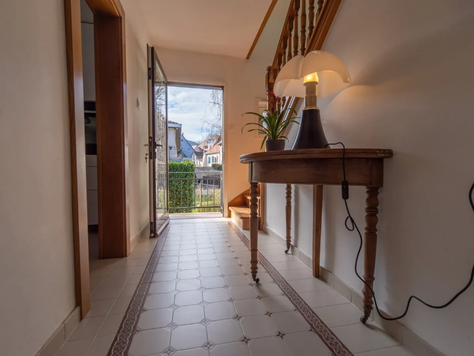 Couloir avec un sol carrelé, une table en bois et une lampe, donnant sur un balcon lumineux.