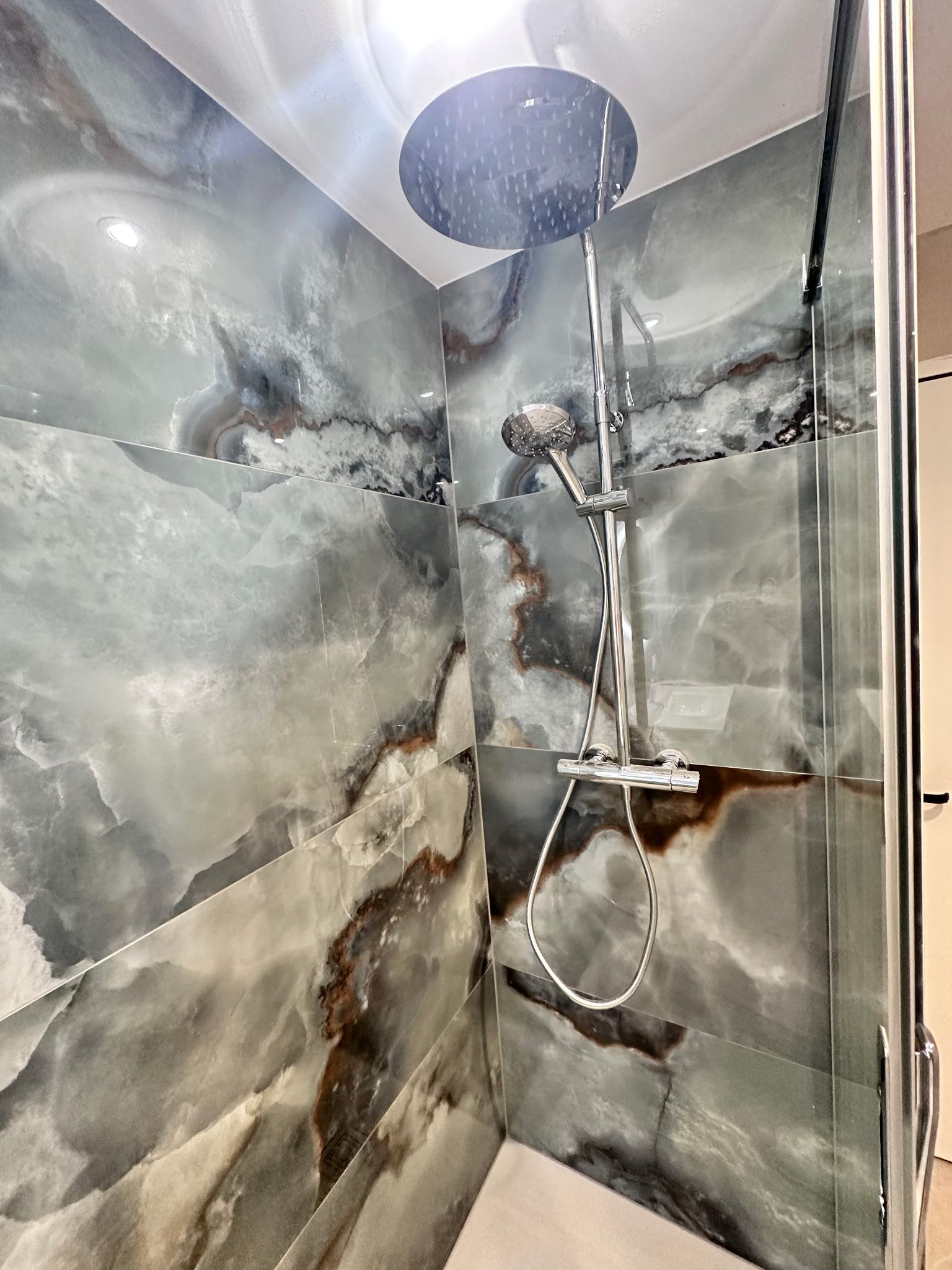 Douche moderne avec du carrelage gris imitation marbre et une douchette chromée dans une cabine d’angle.