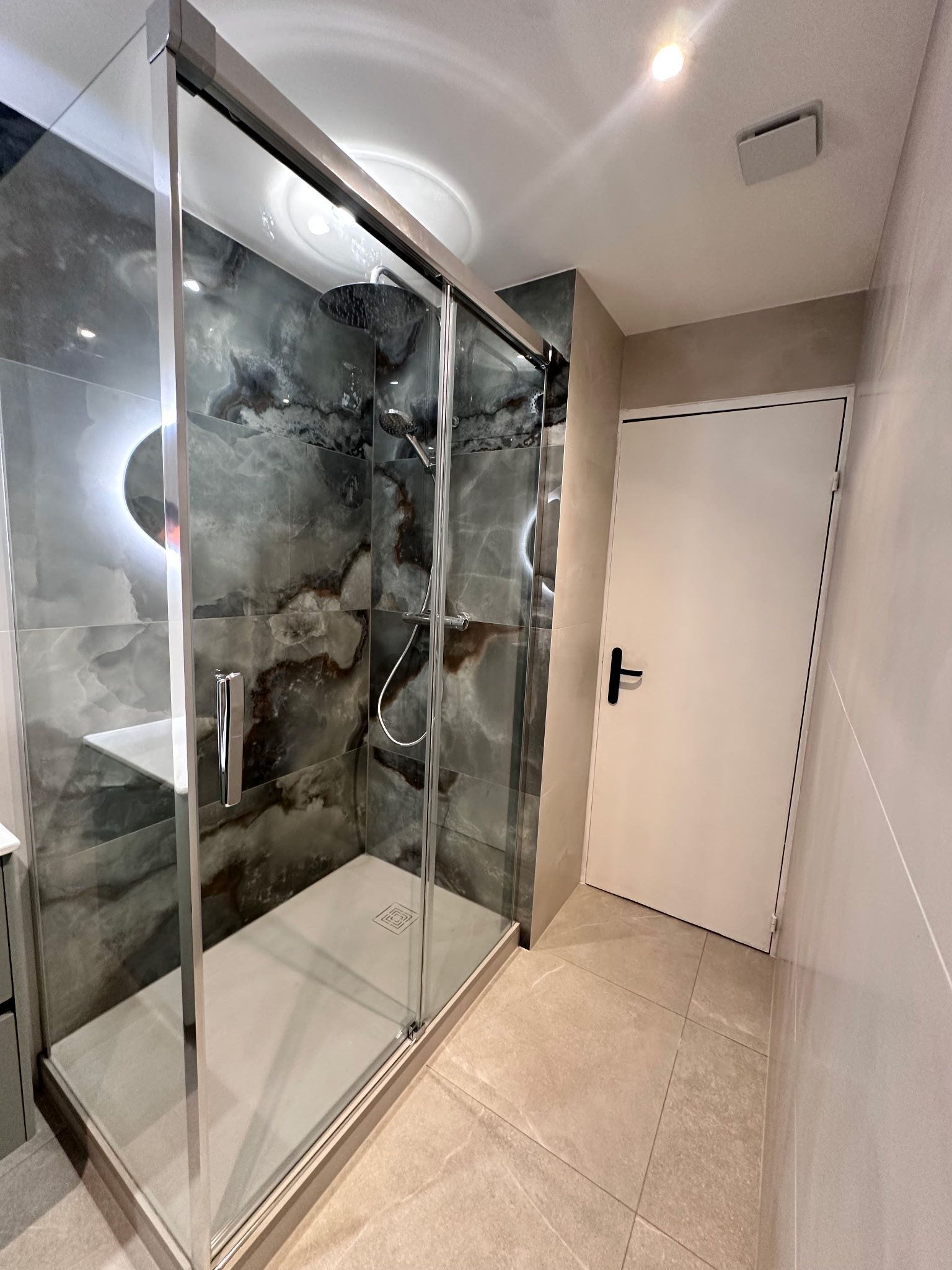 Couloir moderne avec une cabine de douche en verre, un carrelage beige au sol et une porte blanche au fond.