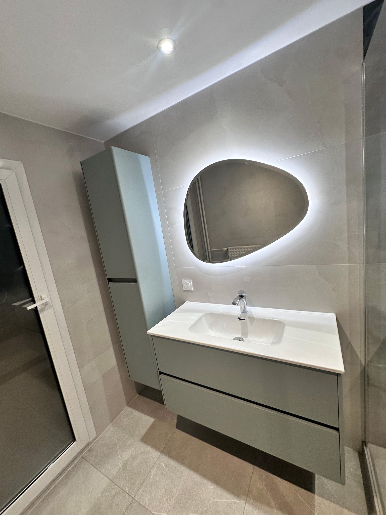Salle de bains moderne avec meuble vasque suspendu, miroir ovale et colonne de rangement sous un éclairage mural lumineux.