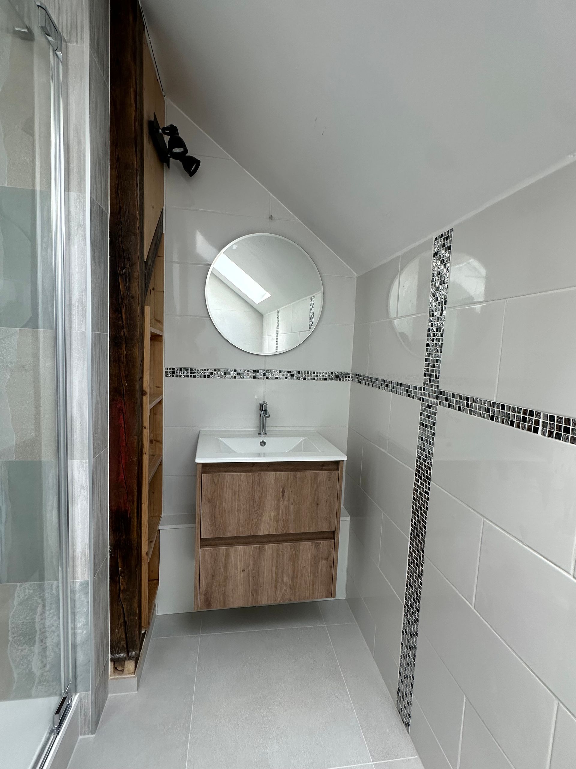 Petite salle de bains moderne avec du carrelage blanc, un miroir rond, un lavabo en bois et une porte de douche en verre.