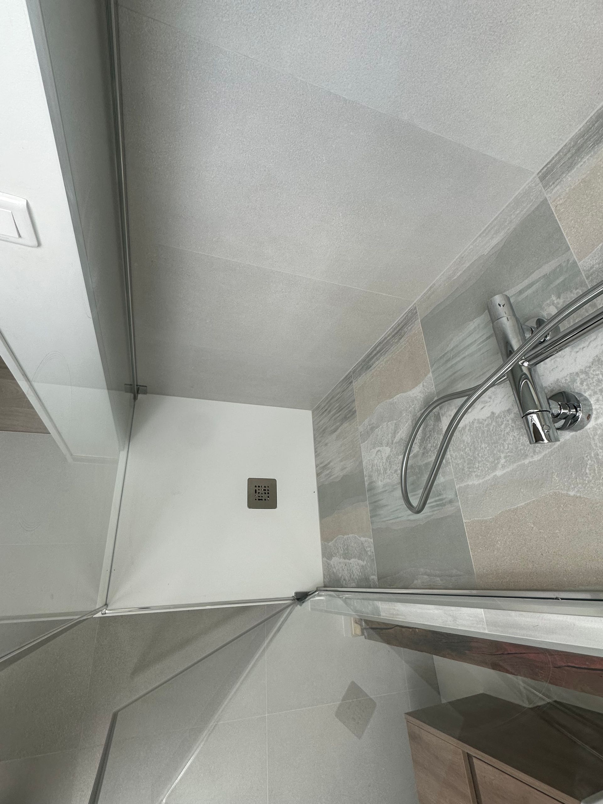 Vue de dessus d’une petite cabine de douche avec du carrelage gris, une porte vitrée et une douchette à main.