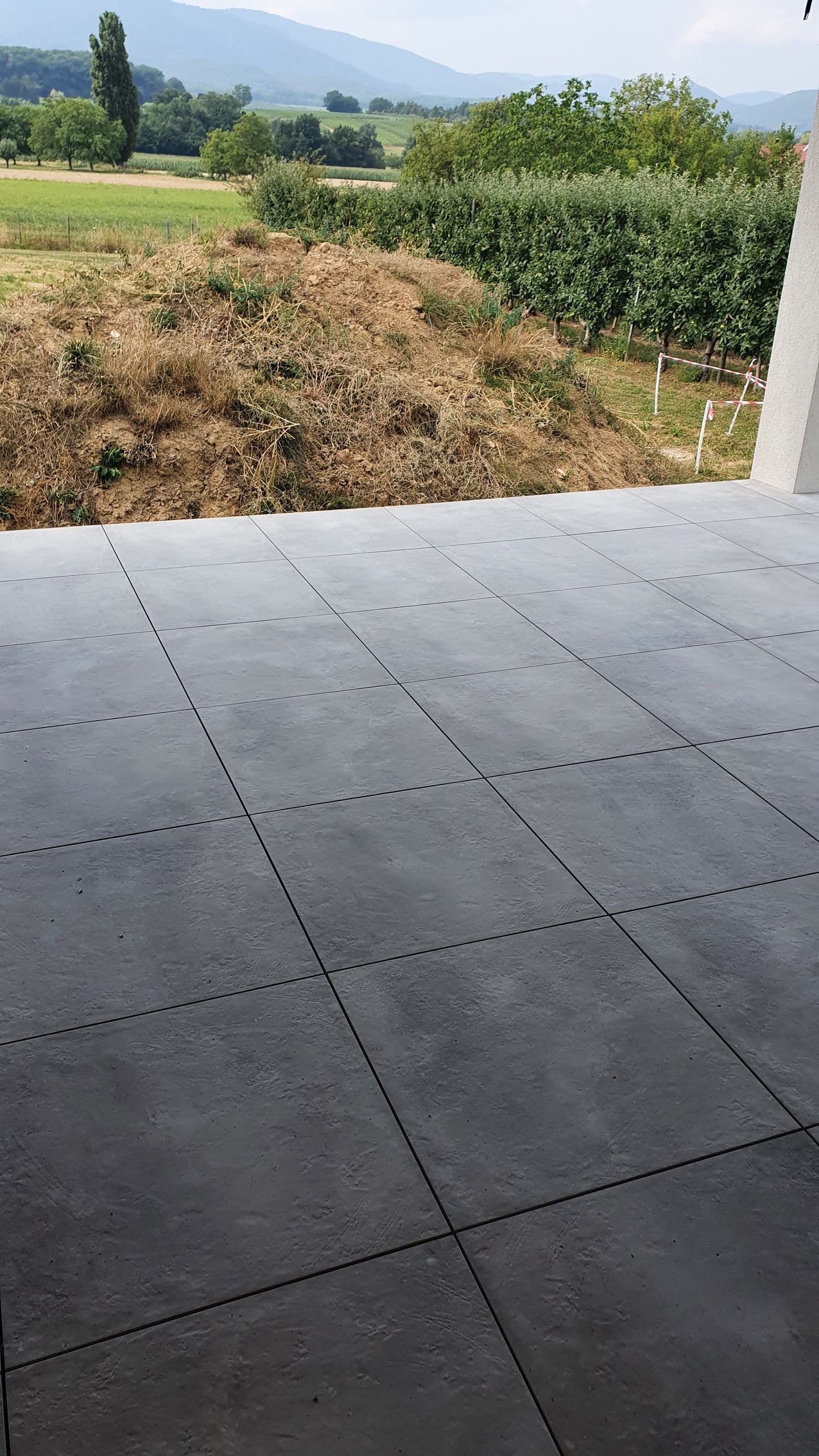 Patio carrelé gris donnant sur un champ verdoyant, un champ de maïs et des collines au loin sous un ciel lumineux