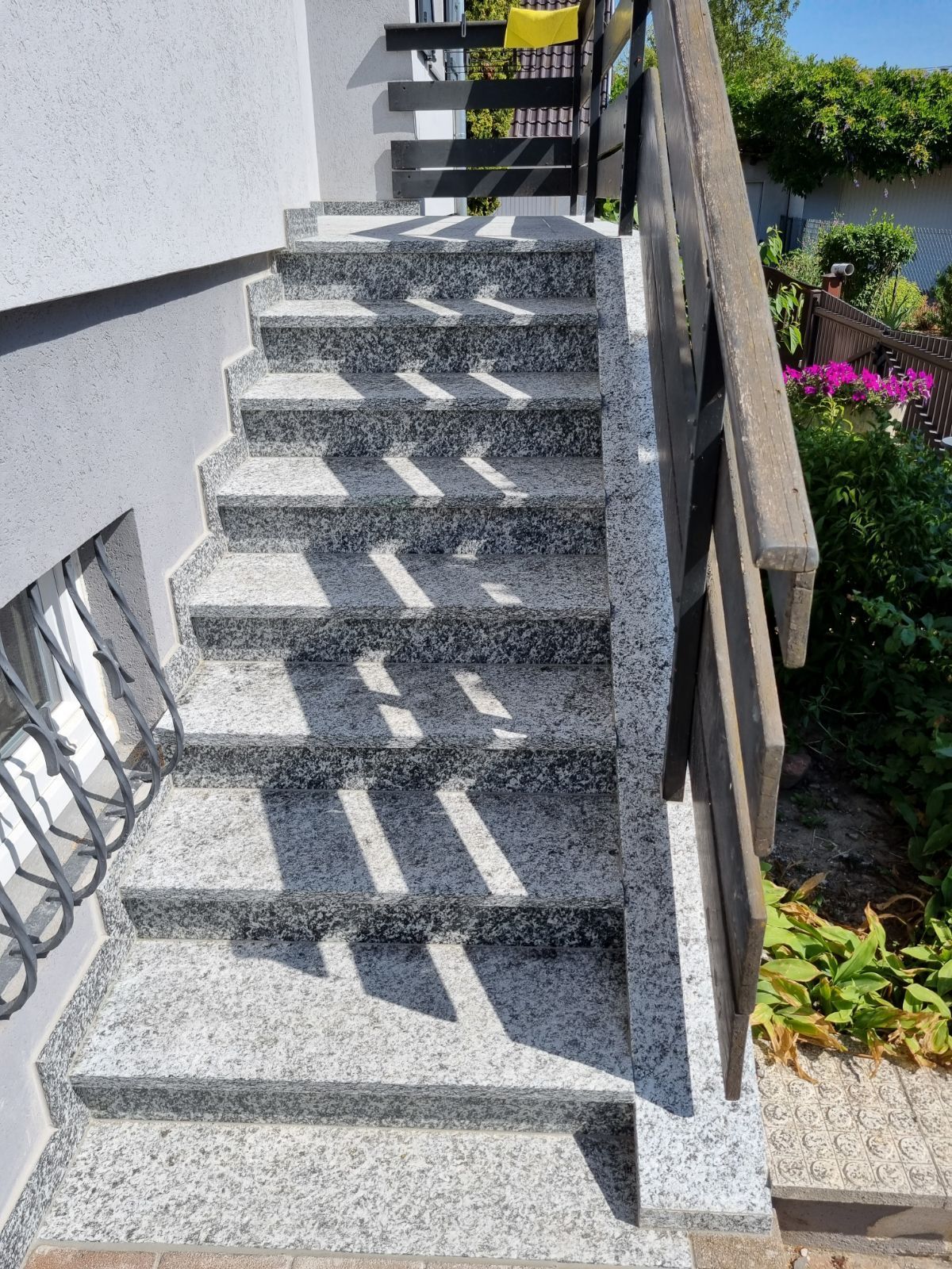 Escalier extérieur en granit longeant un mur blanc et un jardin, avec une rampe en métal qui projette des ombres rayées.
