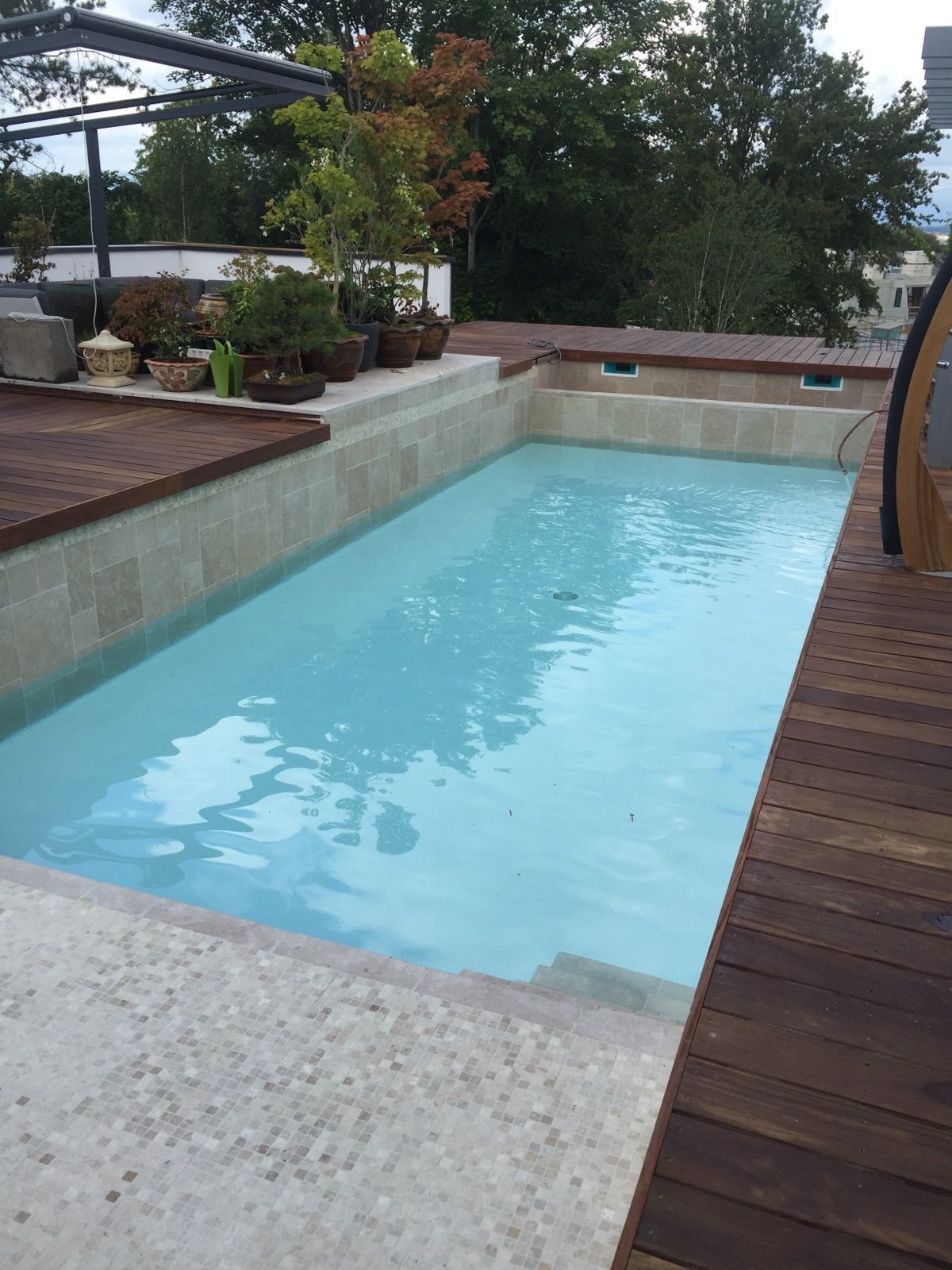 Petite piscine turquoise bordée d’une terrasse en bois et de plantes en pot, dans une cour intérieure