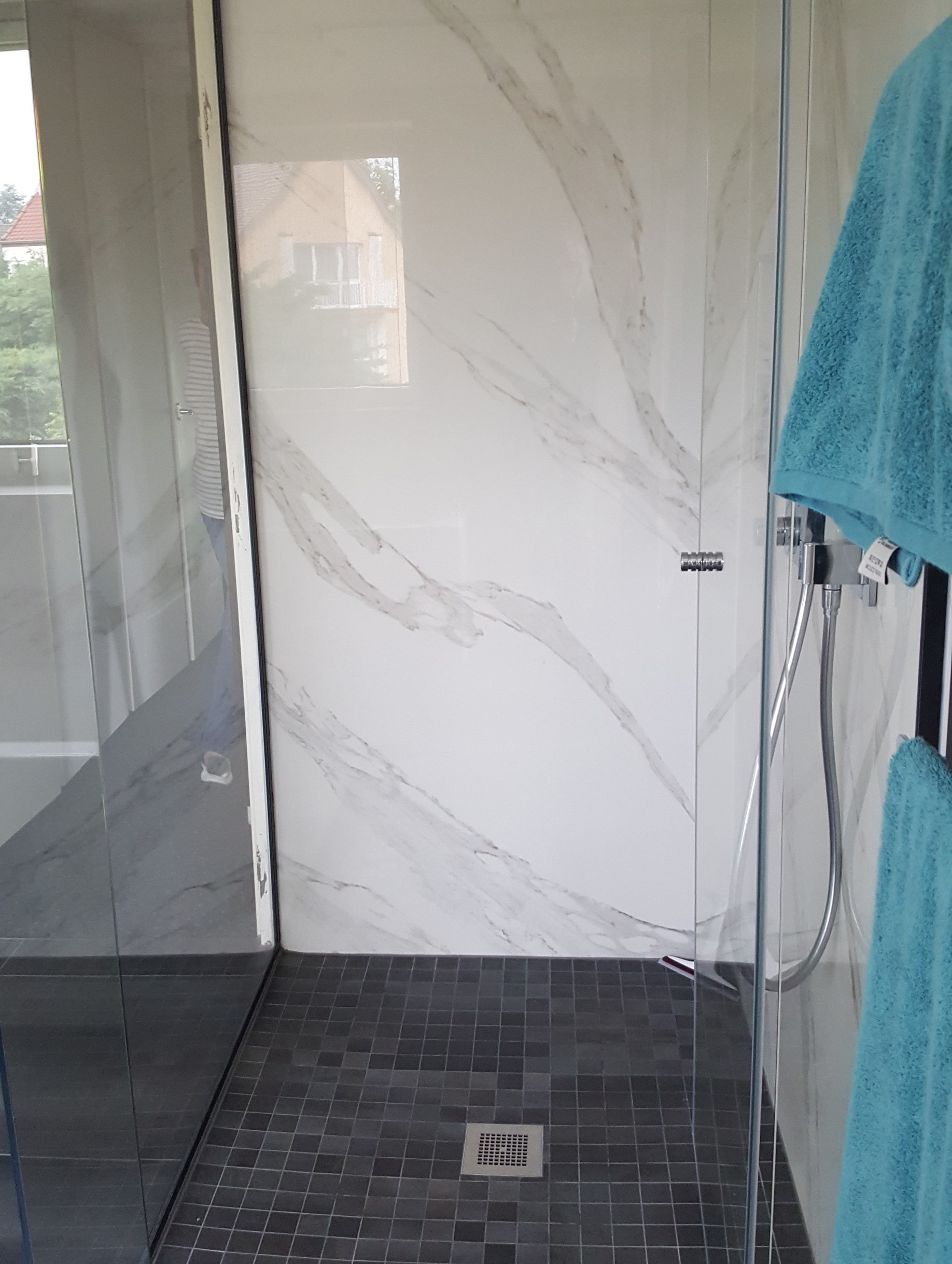 Douche moderne carrelée avec porte en verre, panneau mural imitation marbre blanc et serviette bleue à droite.