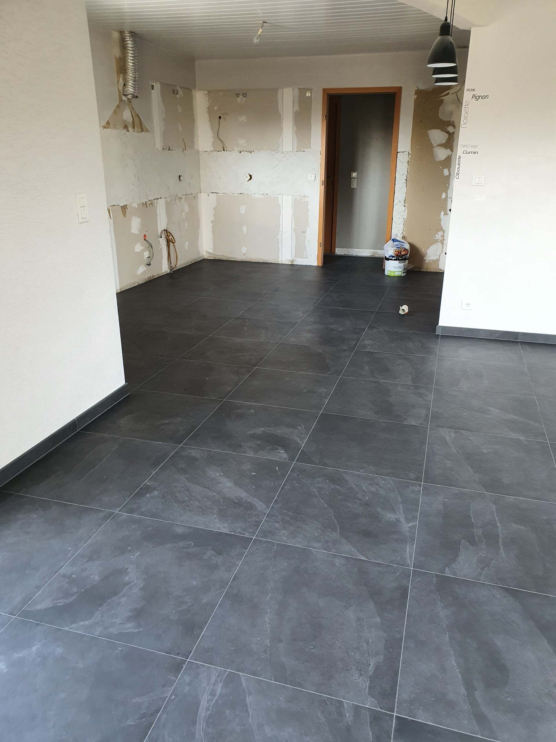 Pièce vide avec un sol carrelé foncé, des murs blancs et une porte au fond.