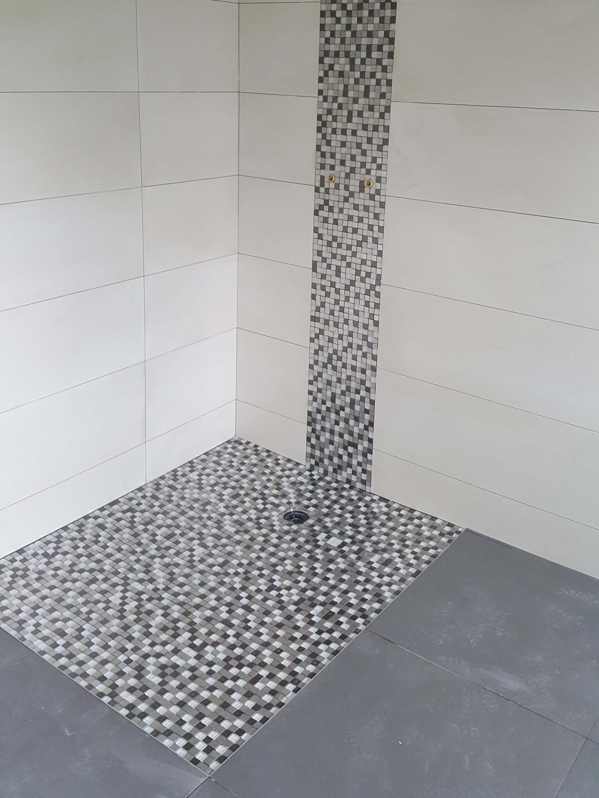 Douche carrelée avec des murs blancs, un sol gris et une bande de mosaïque au sol et au mur.
