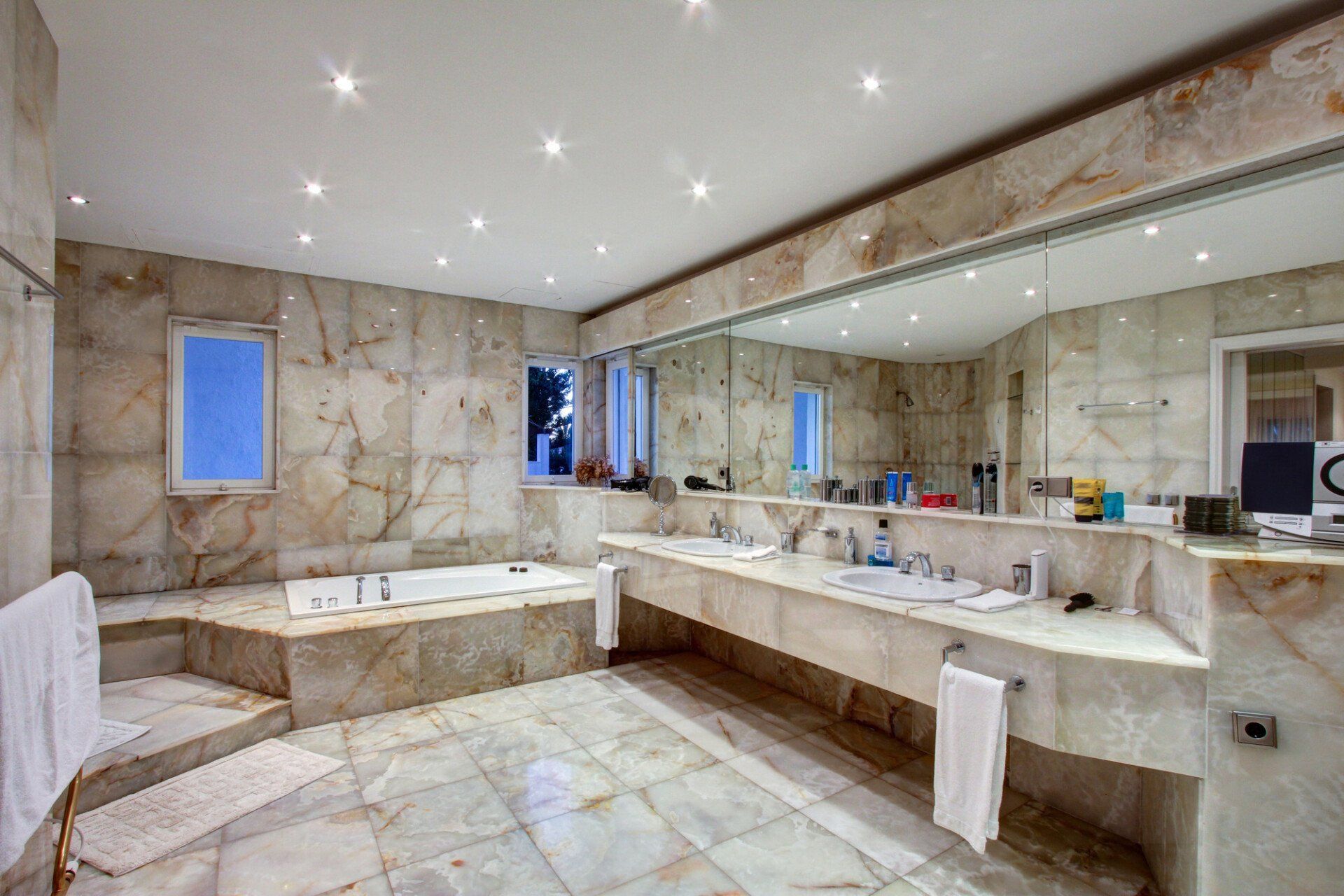 Salle de bains spacieuse en marbre avec double vasque, grand miroir, baignoire et spots encastrés au plafond.